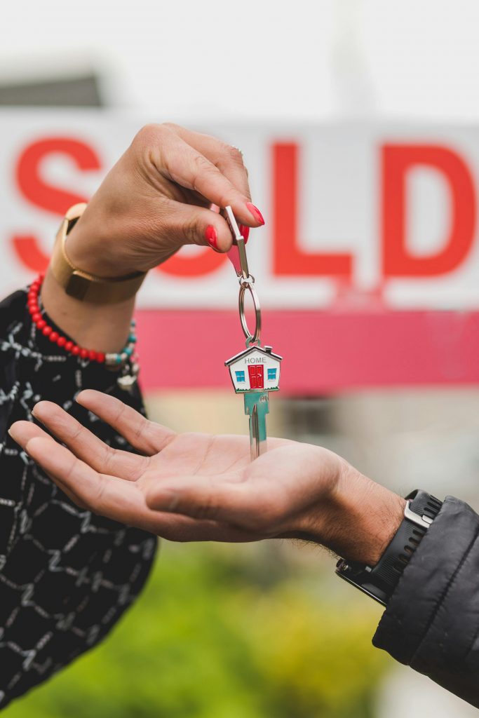 pexels-photo-8293717-8293717 Close-up of hands exchanging house key with colorful SOLD sign in the background.